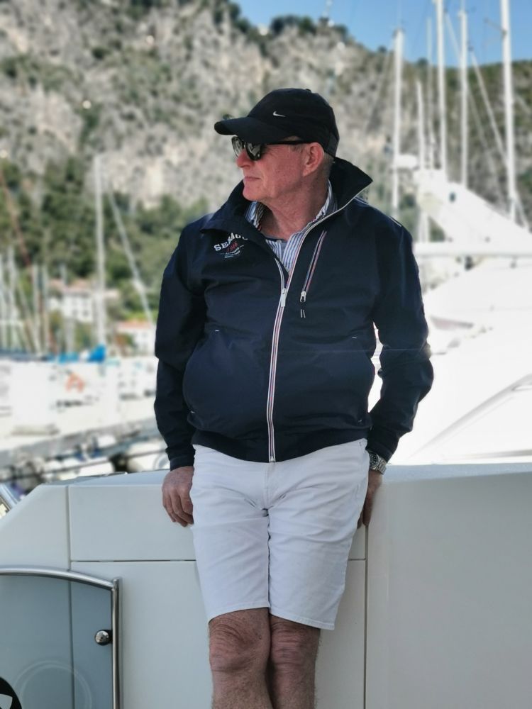 Captain Simon Rees in a blue hat, blue bomber jacket and white shorts looking to his right with boats and french riviera mountains framing the background behind him.