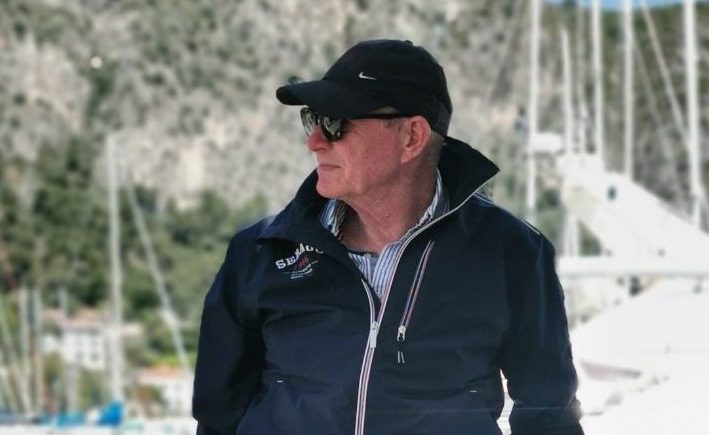 Captain Simon Rees in a blue hat, blue bomber jacket and white shorts looking to his right with boats and french riviera mountains framing the background behind him.